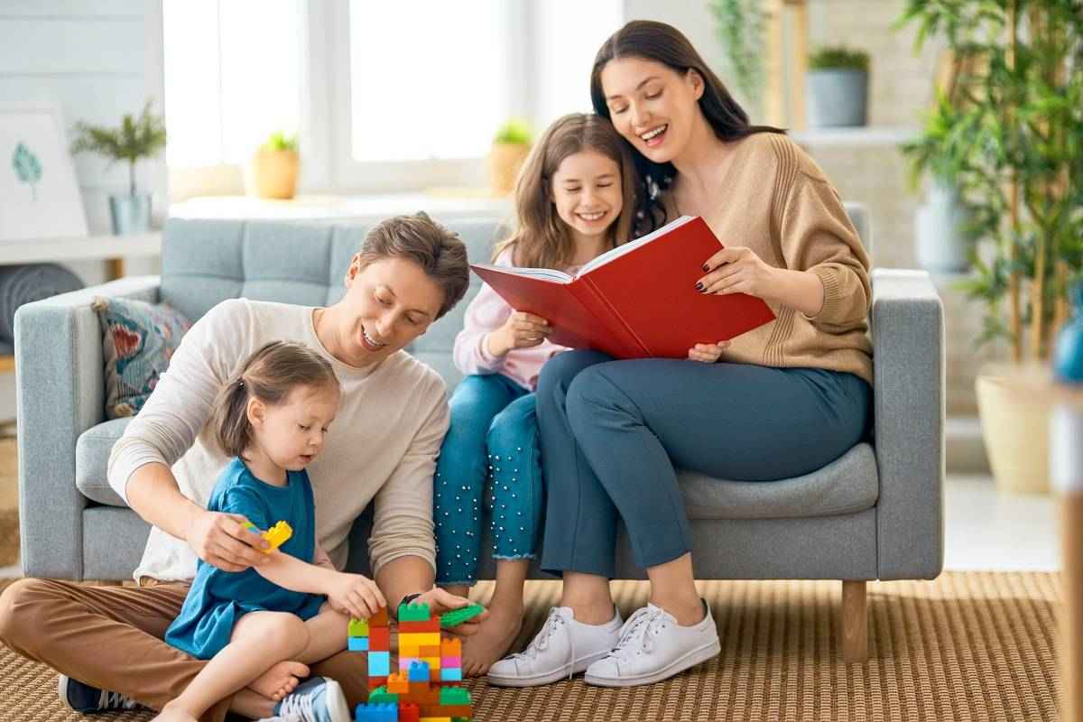  A family is sitting in a living room. The mother is reading a book to the older daughter while the father is playing with blocks with the younger daughter. They are all smiling and appear to be enjoying themselves.